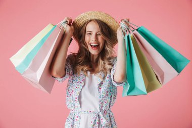 Yazlık elbise ve hasır şapka alışveriş torbaları üzerinde pembe arka plan izole tutan heyecanlı bir genç kadın portresi