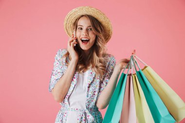 Yazlık elbise ve cep telefonu üzerinde alışveriş torbaları üzerinde pembe arka plan izole tutarken konuşurken hasır şapka neşeli bir genç kadın portresi