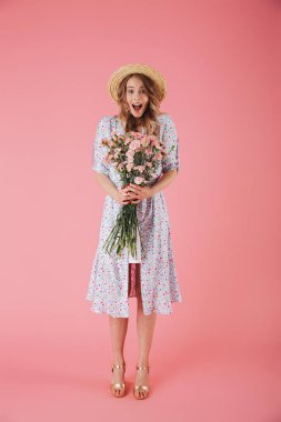 Yazlık elbise ve Karanfil Buketi pembe arka plan üzerinde izole tutarken poz hasır şapka neşeli bir genç kadın portresi tam uzunlukta