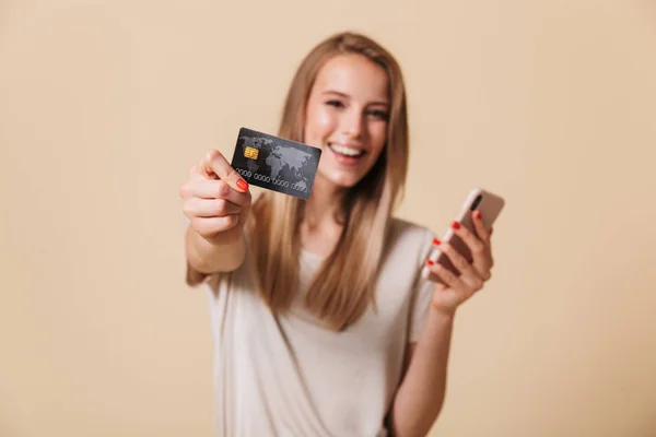 携帯電話とベージュ色の背景に分離されたプラスチック製のクレジット カードを示す幸せなカジュアルな女の子の肖像画 — ストック写真