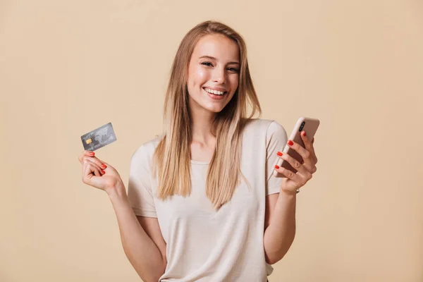 携帯電話とベージュ色の背景に分離されたプラスチック製のクレジット カードを保持している笑顔のカジュアルな女の子の肖像画 — ストック写真