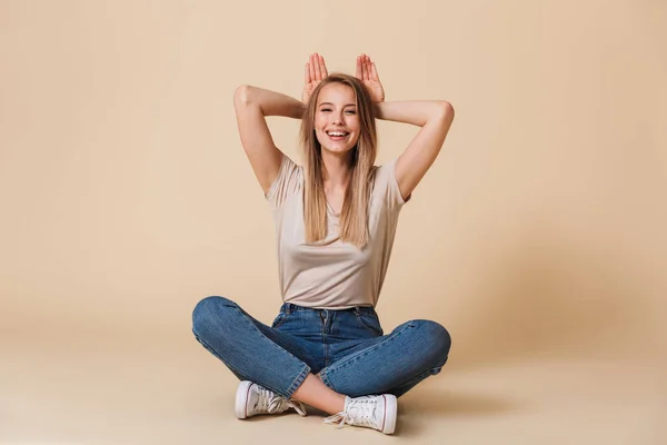 Photo of adorable european woman sitting on floor with legs crossed and showing rabbit ears isolated over beige background