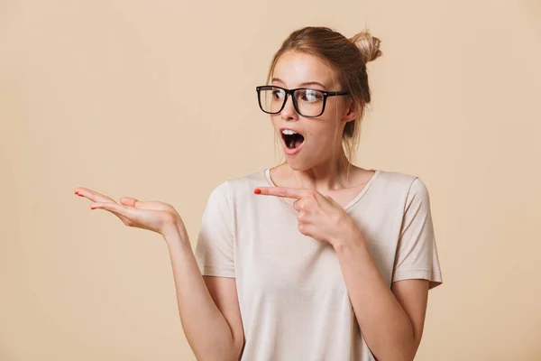 照片特写欧洲妇女与金发绑头发穿着休闲服装和眼镜微笑着 指着手指在 Copyspace 在棕褐色背景下的棕榈隔离 — 图库照片