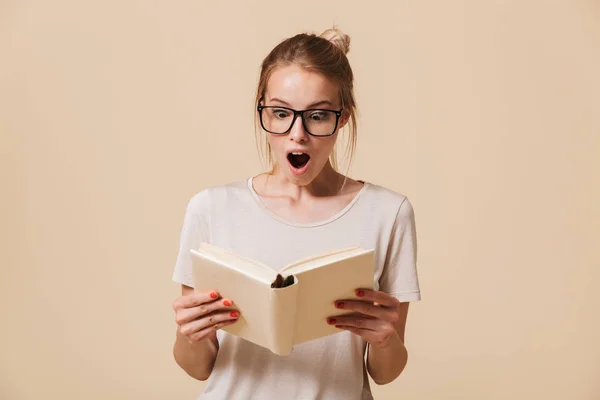 一个震惊的年轻女孩在眼镜的肖像阅读一本书 在米色背景隔离 — 图库照片