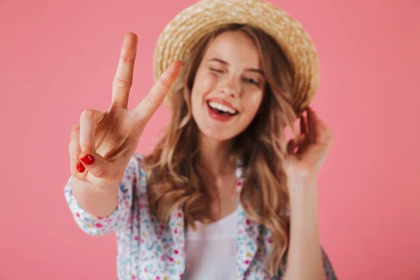 关闭一个微笑的年轻女子在夏季礼服和草帽显示和平姿态隔离粉红色背景的肖像 — 图库照片