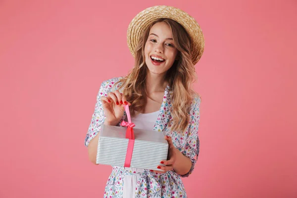 一个快乐的年轻女子在夏季礼服和草帽控股礼品盒在粉红色背景下隔离的肖像 — 图库照片