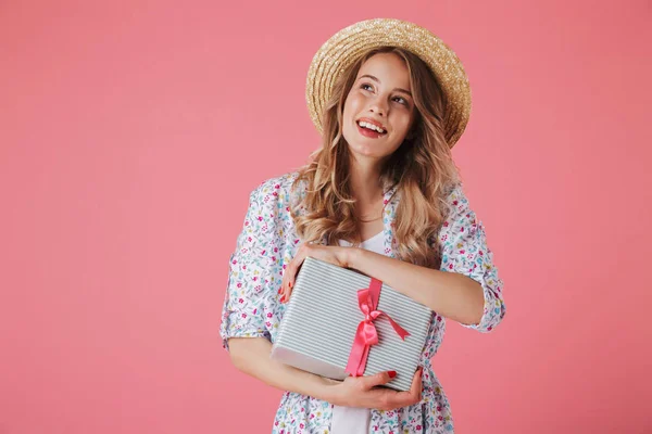 Portrait of a wondering young woman in summer dress and straw hat holding gift box isolated over pink background