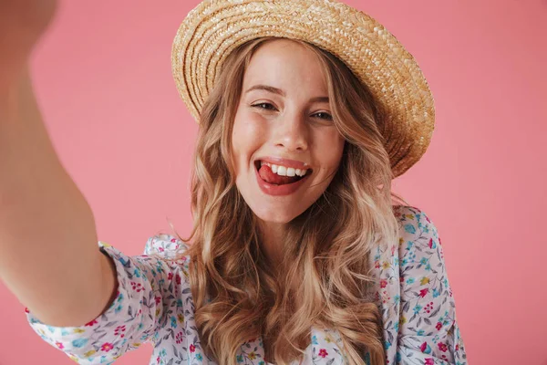 关闭一个顽皮的年轻女子的肖像在夏季礼服和草帽采取自拍孤立的粉红色背景 — 图库照片