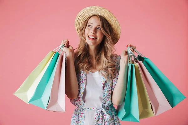 一个年轻快乐的女人在夏天的礼服和草帽拿着购物袋 看着远离粉红色背景的照片 — 图库照片
