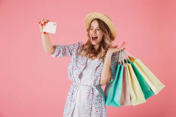 夏のドレスとピンク背景に分離された買い物袋を保持している間 Selfie を取って麦藁帽子で遊び心のある若い女性の肖像画 — ストック写真