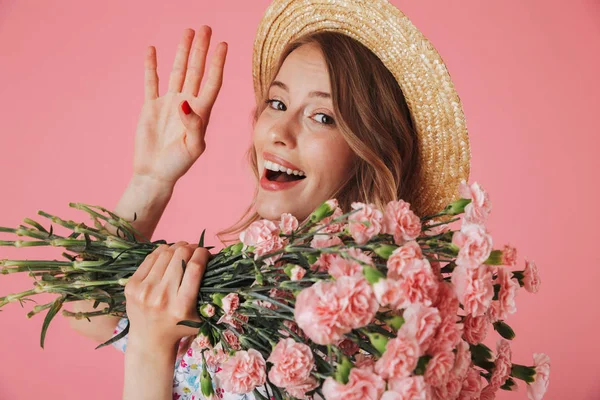 夏のドレスとカーネーションの花束を押しピンク背景に分離の手で振っている麦わら帽子の素敵な若い女性の肖像画間近します — ストック写真