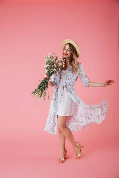 夏のドレスとピンク背景に分離されたカーネーション花束を押しながらポーズ麦わら帽子でかなり若い女性の完全な長さの肖像画 — ストック写真