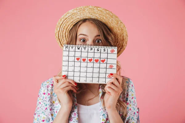 一个漂亮的年轻女子在草帽的肖像持有妇女的期间日历和看着在粉红色背景隔离相机 — 图库照片
