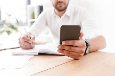 Masada oturan ve onun smartphone--dan parlak office odasında çalışırken not defteri bilgileri aşağı yazma beyaz gömlekli konsantre işadamı resim kırpılmış