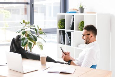 Görüntü office ayaklı masada oturan ve siyah smartphone kullanarak formal giyim mutlu yakışıklı adamın