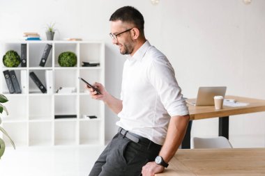 Ofiste tabloda oturma ve iş sohbet için smartphone kullanarak beyaz gömlek giymiş yakışıklı neşeli adam