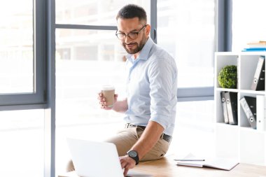 Ciddi adam ofiste tabloda oturma ve çalışma sırasında paket servisi olan restoran kahve içmek için gümüş dizüstü kullanarak beyaz gömlek giymiş görüntüsünü