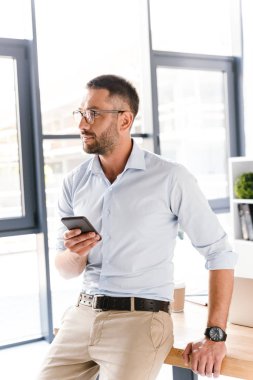 İcra Müdürü adam şık giyim ve ofiste tabloda oturan ve cep telefonu kullanırken büyük pencereden bakarak gözlük görüntüsünü