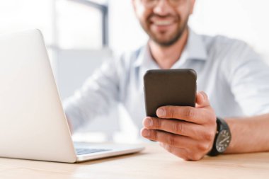 Yetişkin Erkek 30'lu yaşlarda beyaz gömlekli ise masada oturan ve ofiste çalışan dizüstü bilgisayar ve cep telefonu kullanarak gülümseyen fotoğraf kırpılmış
