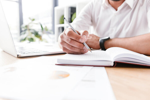 Cropped image of adult business man in white shirt sitting at table and writing down information with pencil at notebook while working in bright office room