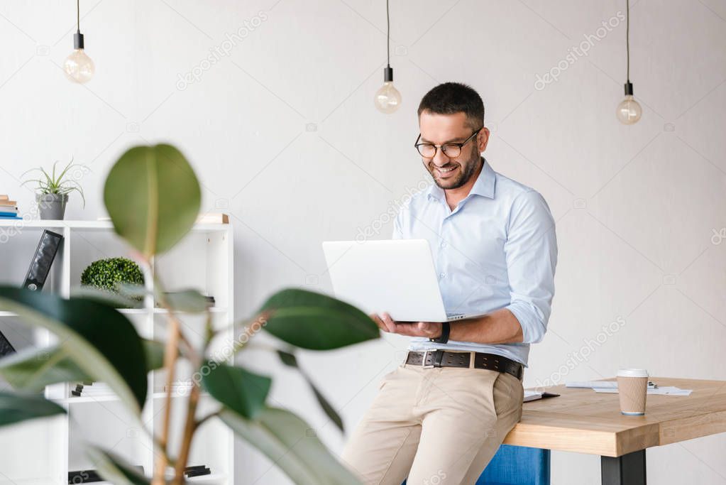 Smiling satisfied man 30s wearing white shirt sitting on table in office and having business chat on silver laptop