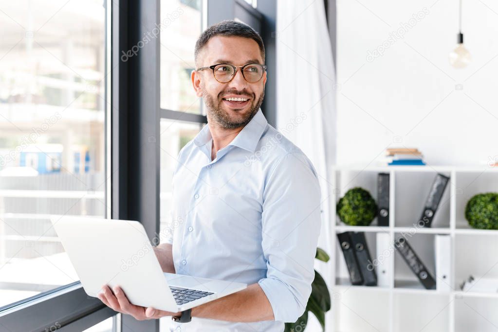 Image of office worker man wearing white shirt looking aside while holding silver laptop during work in business centre