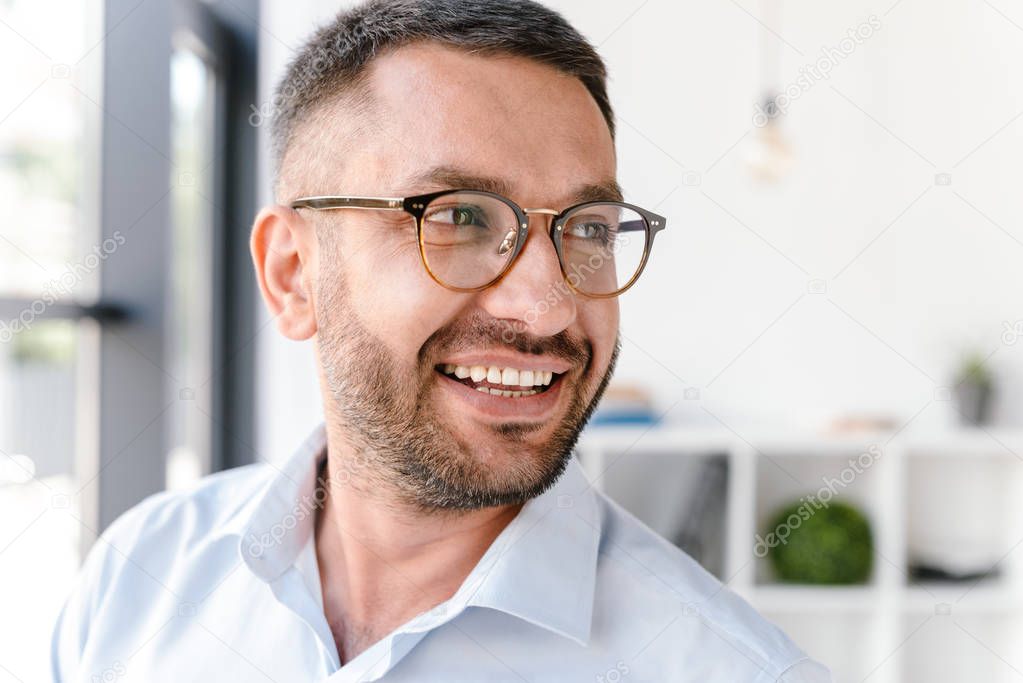 Portrait closeup of handsome man 30s wearing white shirt and glasses looking aside while standing near big window in business centre or office room
