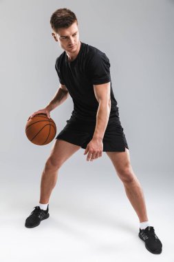Full length portrait of a confident young sportsman playing basketball isolated over gray background