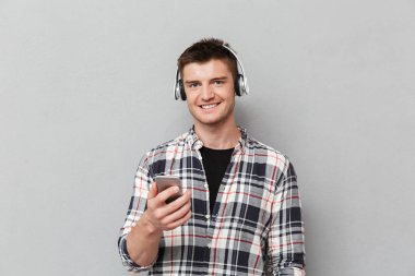 Kulaklık ile müzik dinlemek ve cep telefonu gri arka plan üzerinde tutarak gülümseyen genç bir adam portresi