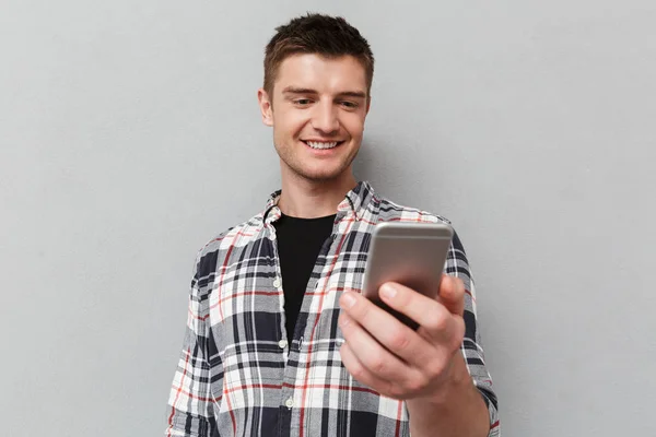 一个微笑的年轻人在灰色背景下看手机的肖像 — 图库照片