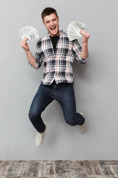 灰色の背景を飛び越えながらお金紙幣を示す陽気な若い男の完全な長さの肖像画 — ストック写真