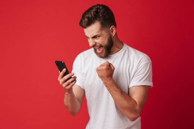Genç adam cep telefonuyla kırmızı duvar arka plan üzerinde izole ayakta azarlama görüntü kazanan jest yapmak.