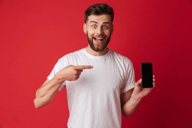 Heyecanlı genç adam cep telefonu işaret ekran gösterilen Kırmızı duvar arka plan üzerinde izole ayakta görüntüsünü.