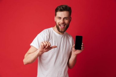 Heyecanlı genç adam cep telefonu işaret ekran gösterilen Kırmızı duvar arka plan üzerinde izole ayakta görüntüsünü.
