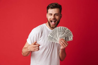 Heyecanlı genç adam para işaret tutan kırmızı duvar arka plan üzerinde izole görüntüsünü.