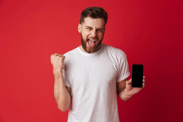 携帯電話のディスプレイを示す赤い壁背景に分離立って興奮して若い男のイメージを作る勝者ジェスチャー — ストック写真