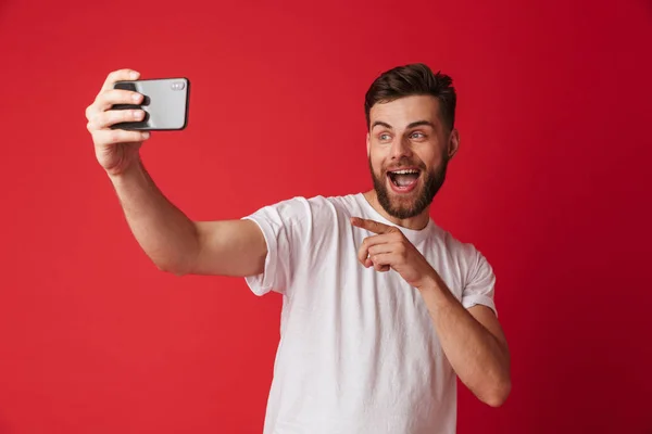 携帯電話を指すことによって分離赤い壁背景を Selfie の上に立っている感情的な若い男のイメージ — ストック写真