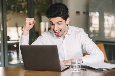 Fotoğraf heyecanlı genç adam 30'lu yaşlarda beyaz gömlek giyiyor ve çığlık ve laptop Office bakarken kazanan gibi yumruk kenetlenmesi bluetooth kulaklıkları