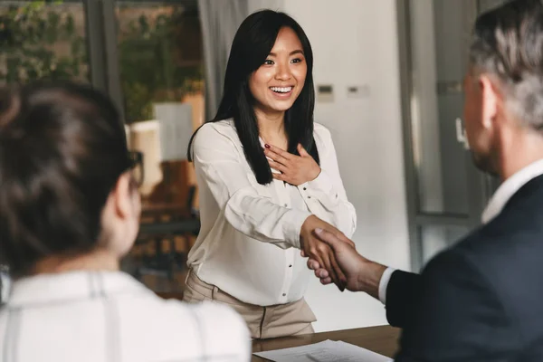 Business, career and placement concept - two business partners in office shaking hand of young asian woman after successful negotiations or interview