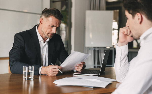 Business, career and placement concept - caucasian businessman negotiating with male candidate about work while reading his resume during job interview in office