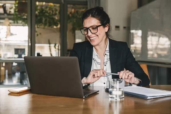 Beyaz patronu kadın 30'lu giyen resmi giyim ve gözlük gülme ya da ofiste masada oturan ve dizüstü bilgisayarınızın ekranına bakarak gülen fotoğrafı