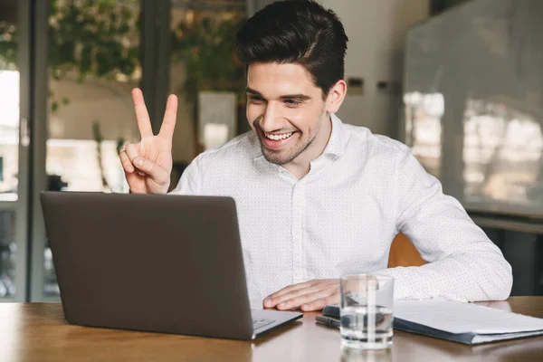 Image of joyful office man 30s wearing white shirt laughing and showing peace sign at laptop during video conference or call