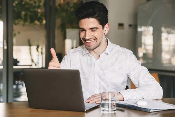 成功商人的肖像30S 穿着白衬衫笑着 在办公室的笔记本电脑上显示拇指在视频会议或电话使用蓝牙耳塞 — 图库照片
