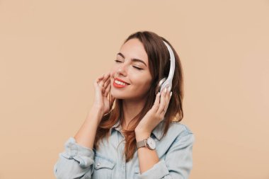 Genç görüntü mutlu kadın gözleri ile bej arka duvarın üzerinden izole müzik dinlerken kulaklık kapalı memnun.
