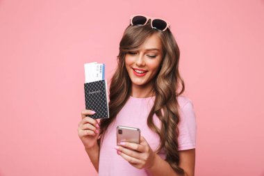 Görüntü bir yolculuk kız pembe arka plan üzerinde kahverengi kilitleri pasaport ve hava bilet tutarken smartphone kullanarak izole güzel olan 20'li