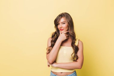 Fotoğraf gülümseyen ve kurnaz bakışla sarı arka plan üzerinde izole çenesinin dokunmadan rahat giysiler giyiyor uzun kahverengi saçlı Genç kadının 20'li