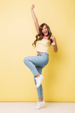 Full length photo of pleased woman 20s with long brown hair rejoicing and screaming in delight with clenching fists isolated over yellow background clipart