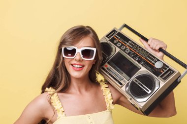 Gülümseyen bir genç kız yaz giysileri ve retro pikap izole sarı arka plan üzerinde tutan güneş gözlüğü portresi