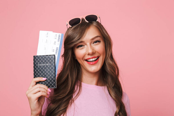 Image of european woman having beautiful brown locks smiling while holding passport and air tickets isolated over pink background