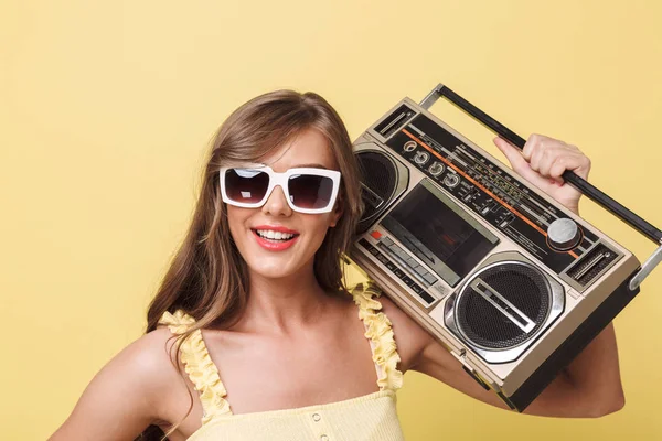 Gülümseyen bir genç kız yaz giysileri ve retro pikap izole sarı arka plan üzerinde tutan güneş gözlüğü portresi